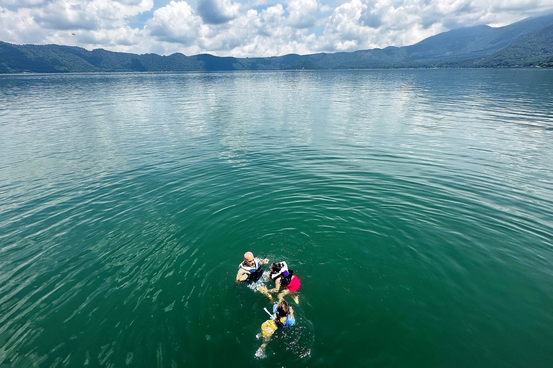 Nuestra familia nadando con chalecos salvavidas en el agua verde del lago de Coatepeque, con el borde del cráter y nubes al fondo